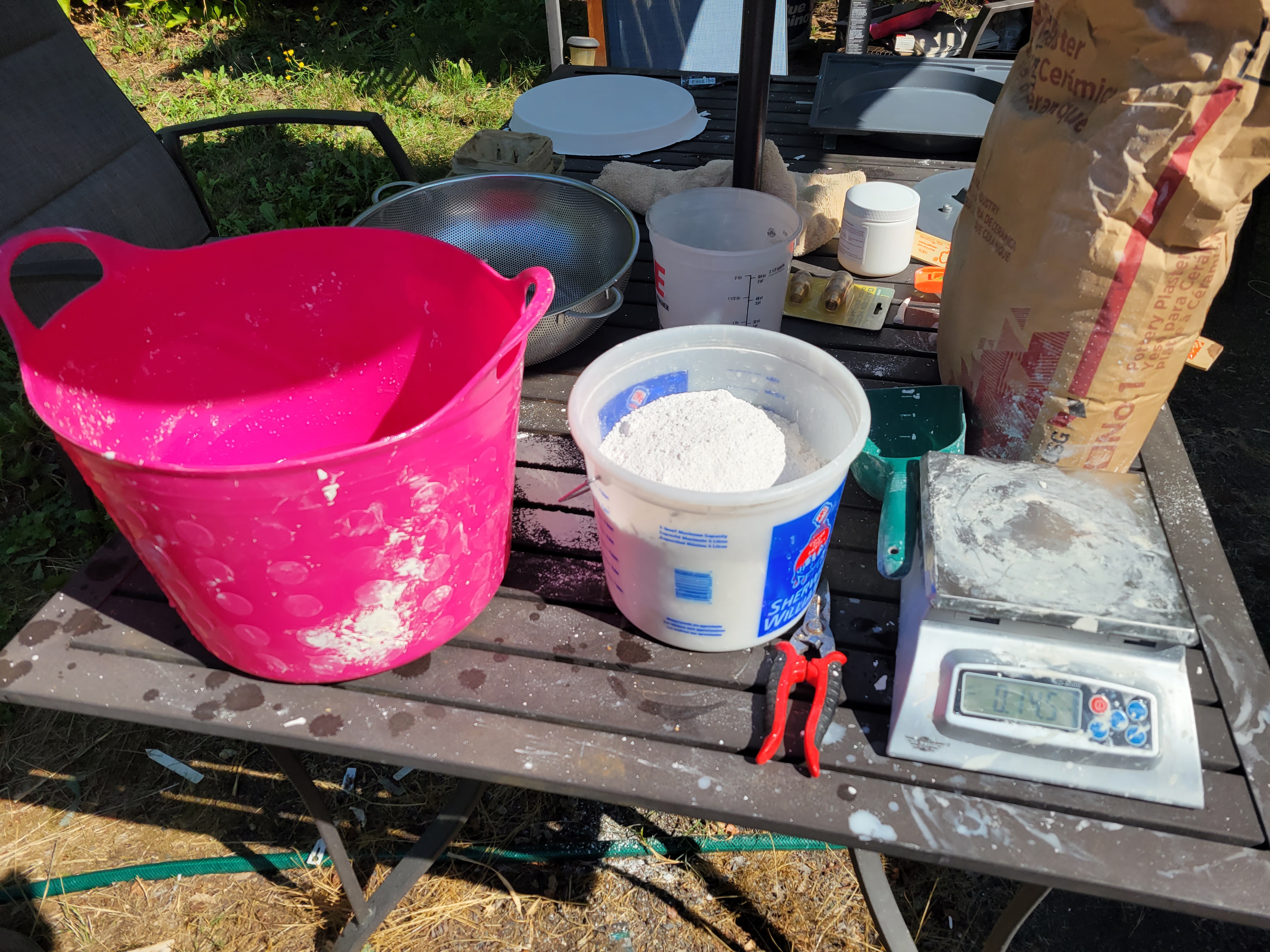 incomplete set of equipment for making plaster bats, including flexible feed bucket, plastic measuring bucket full of powdered plaster, large bag of usg#1 plaster, plastic scoop, and digital scale