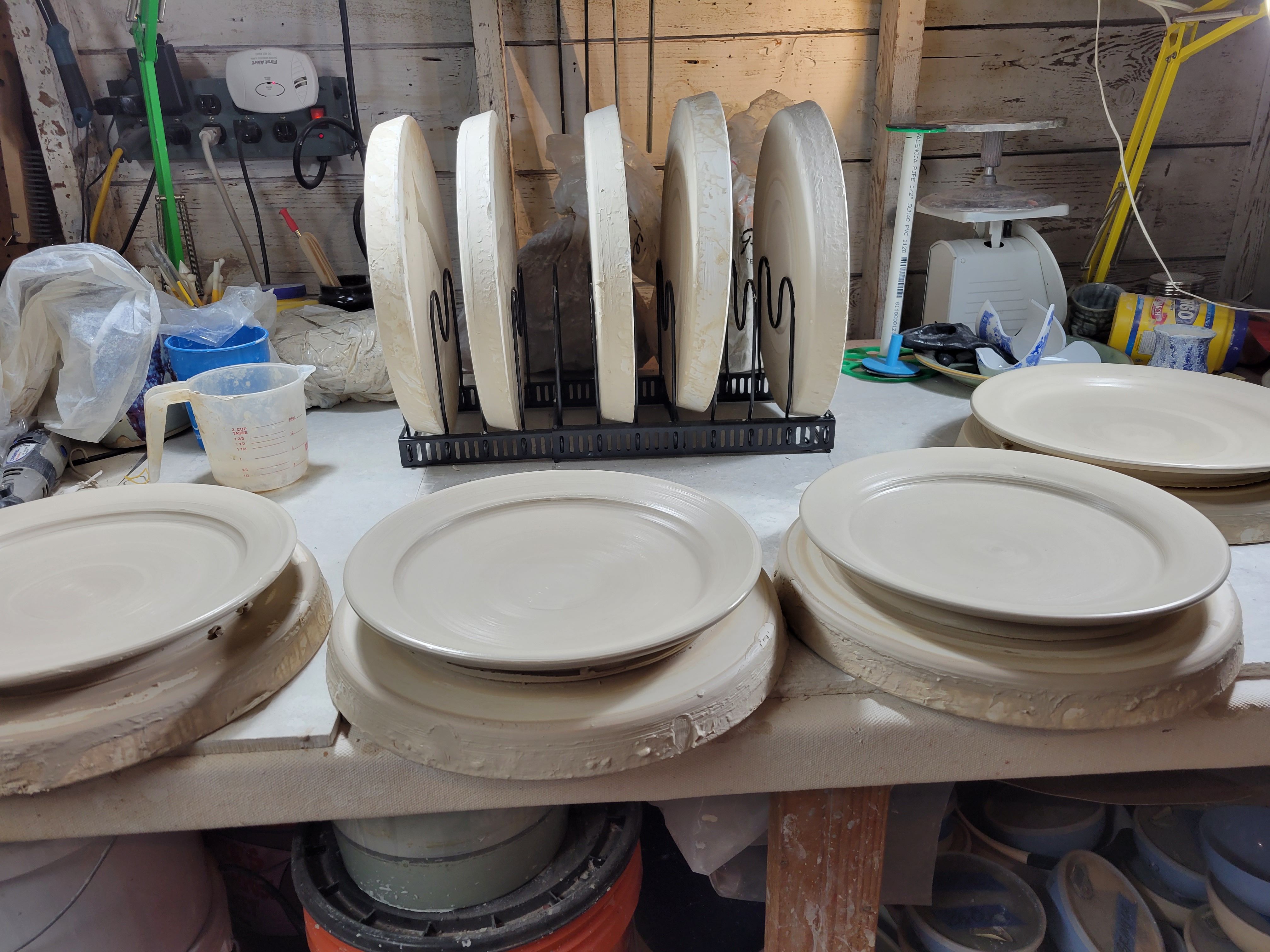 my studio worktable, holding four plates drying on plaster bats.  in the background are five more plaster bats in a metal rack, plus various tools